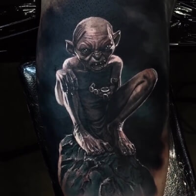 A very dark black and grey tattoo by J Becerra with Gollum sat on a rock, holding the One Ring.