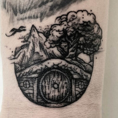 A blackwork tattoo by Adrian de Largue showing a Hobbit House with the Lonely Mountains, Smaug and a tree in the background.