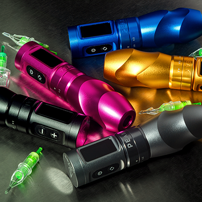 A collection of 5 rotary tattoo machines in black, grey, pink, orange, and blue, all spread of a table with tattoo cartridges next to them.